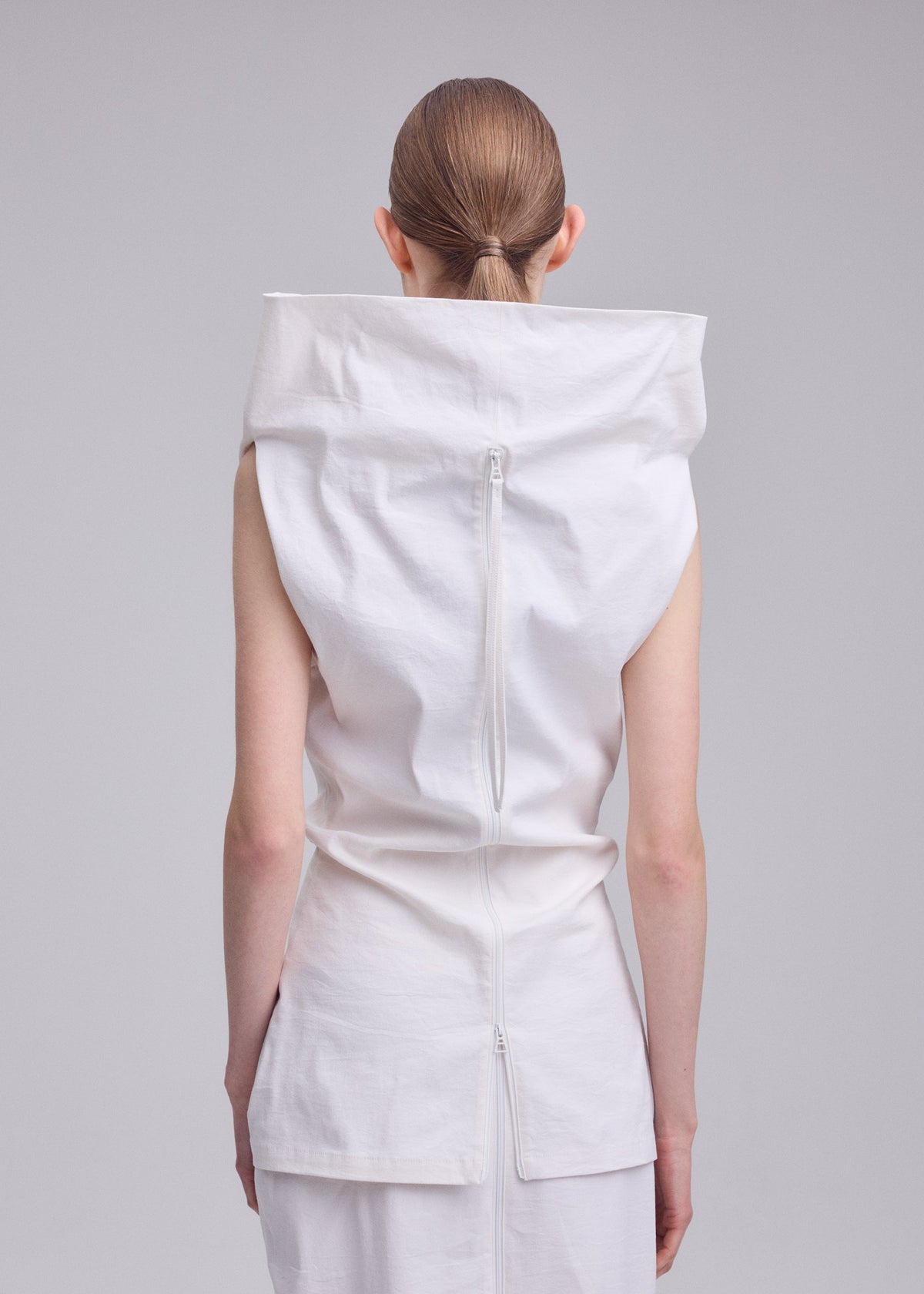 A person with brown hair tied back is shown from behind wearing the ALLOW top by ISSEY MIYAKE, featuring white washi paper-blend fabric, a high collar, and a long vertical back zipper against a light gray background. view 8 of 8