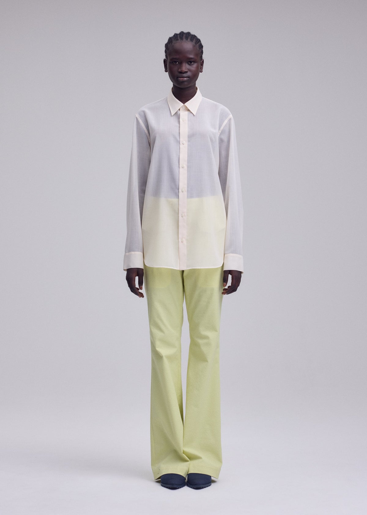 A person stands against a plain light background, dressed in ISSEY MIYAKE's SHEER WOOL shirt in white and pale yellow, paired with light green wide-leg pants and dark shoes. view 5 of 7