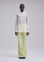 A person stands against a plain light background, dressed in ISSEY MIYAKE's SHEER WOOL shirt in white and pale yellow, paired with light green wide-leg pants and dark shoes.
