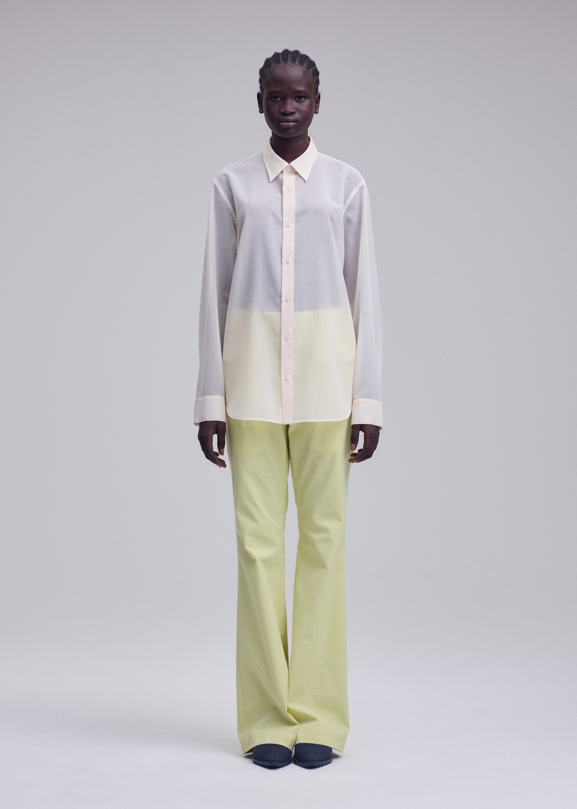 Zoomed view A person stands against a plain light background, dressed in ISSEY MIYAKE's SHEER WOOL shirt in white and pale yellow, paired with light green wide-leg pants and dark shoes.