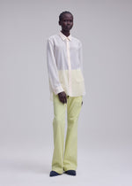 A person stands in a minimal studio wearing the ISSEY MIYAKE SHEER WOOL loose, cream and pale yellow color-block washable wool shirt with light yellow pants and black shoes against a plain light gray background.