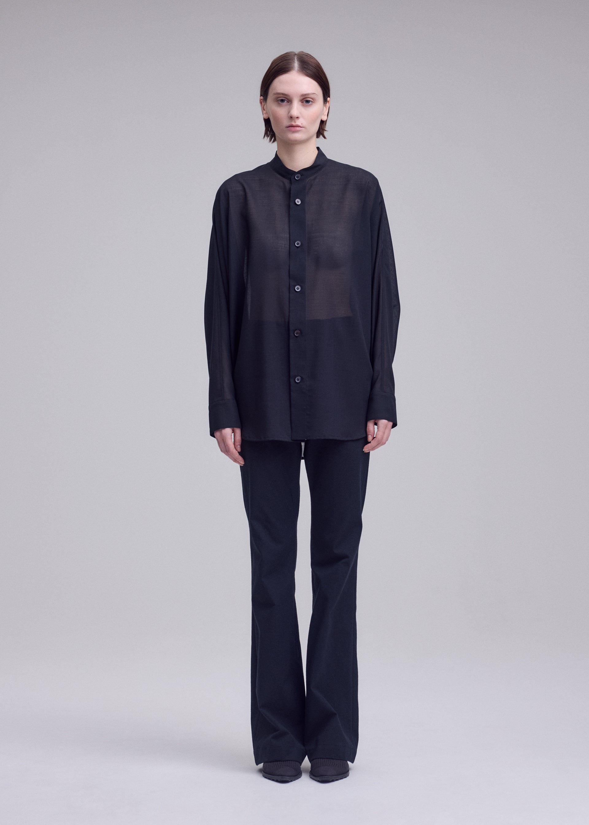 Zoomed view A person faces forward against a plain background, wearing ISSEY MIYAKE's SHEER WOOL: a loose, black semi-sheer shirt paired with black pants and shoes. Their arms rest relaxed at their sides.