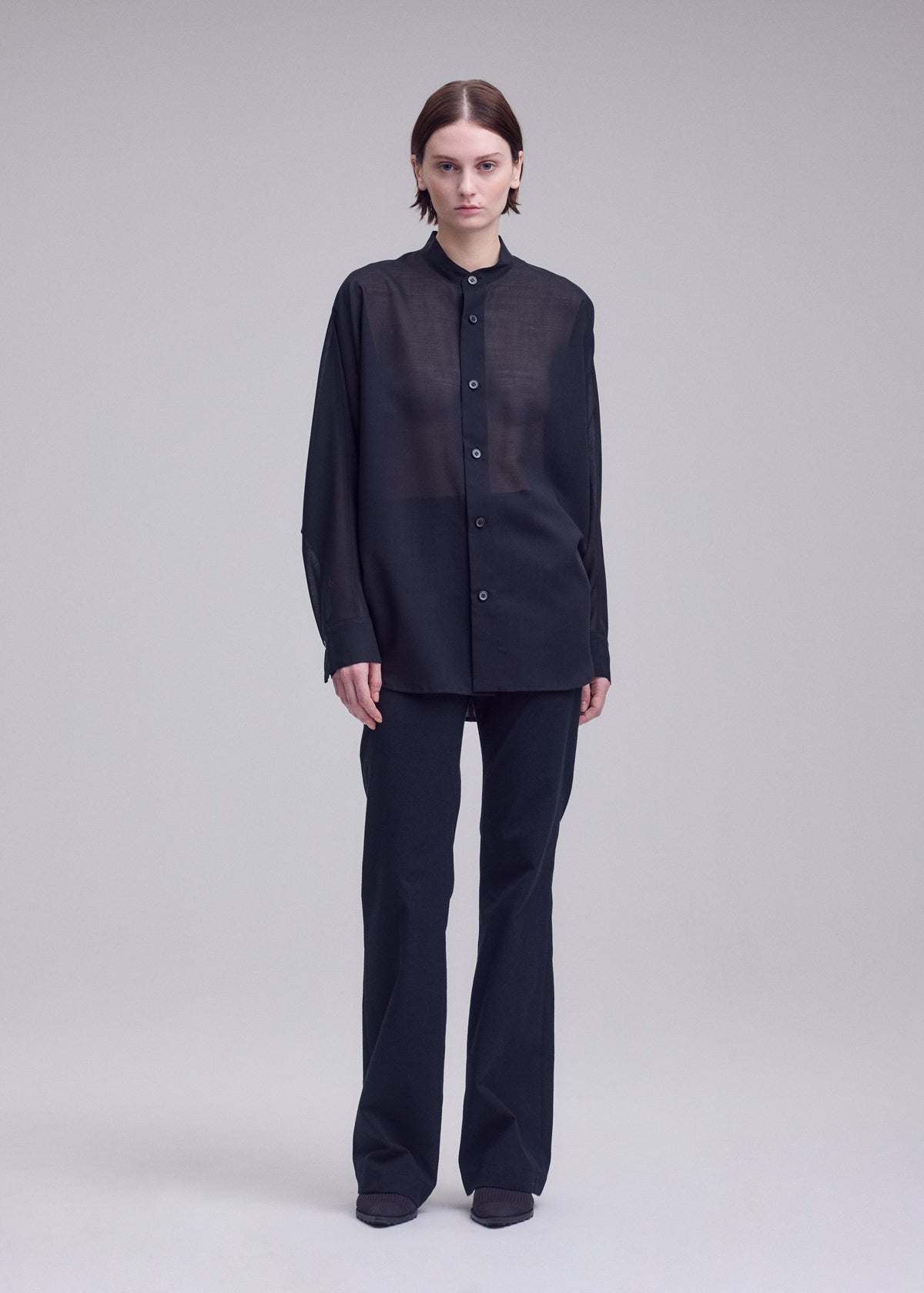 A person with short brown hair stands against a plain light background, wearing ISSEY MIYAKE's SHEER WOOL loose-fitting, long-sleeved button-up shirt and straight black pants paired with black shoes. view 6 of 8