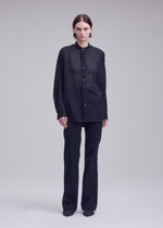 A person with short brown hair stands against a plain light background, wearing ISSEY MIYAKE's SHEER WOOL loose-fitting, long-sleeved button-up shirt and straight black pants paired with black shoes.