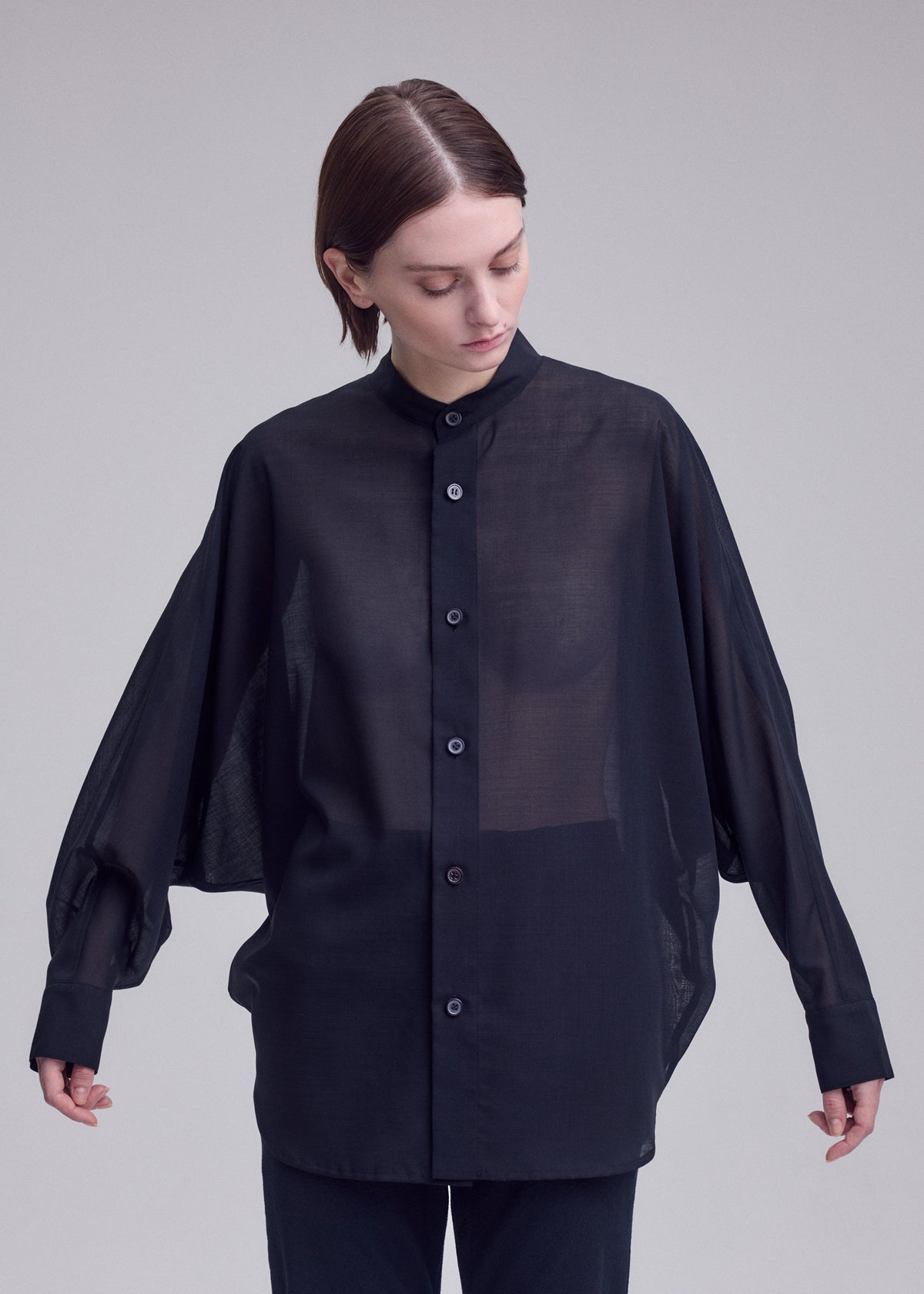 A person with short brown hair wears ISSEY MIYAKE's SHEER WOOL, a loose black dolman-sleeve shirt with long sleeves and black pants, standing against a plain light gray background and looking down. view 7 of 8