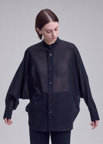 A person with short brown hair wears ISSEY MIYAKE's SHEER WOOL, a loose black dolman-sleeve shirt with long sleeves and black pants, standing against a plain light gray background and looking down.