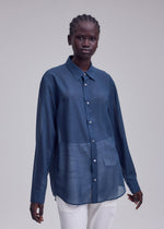 A person with dark skin and braided hair models the ISSEY MIYAKE SHEER WOOL, a loose-fitting, long-sleeved, button-up blue shirt with white pants, against a plain light gray background.