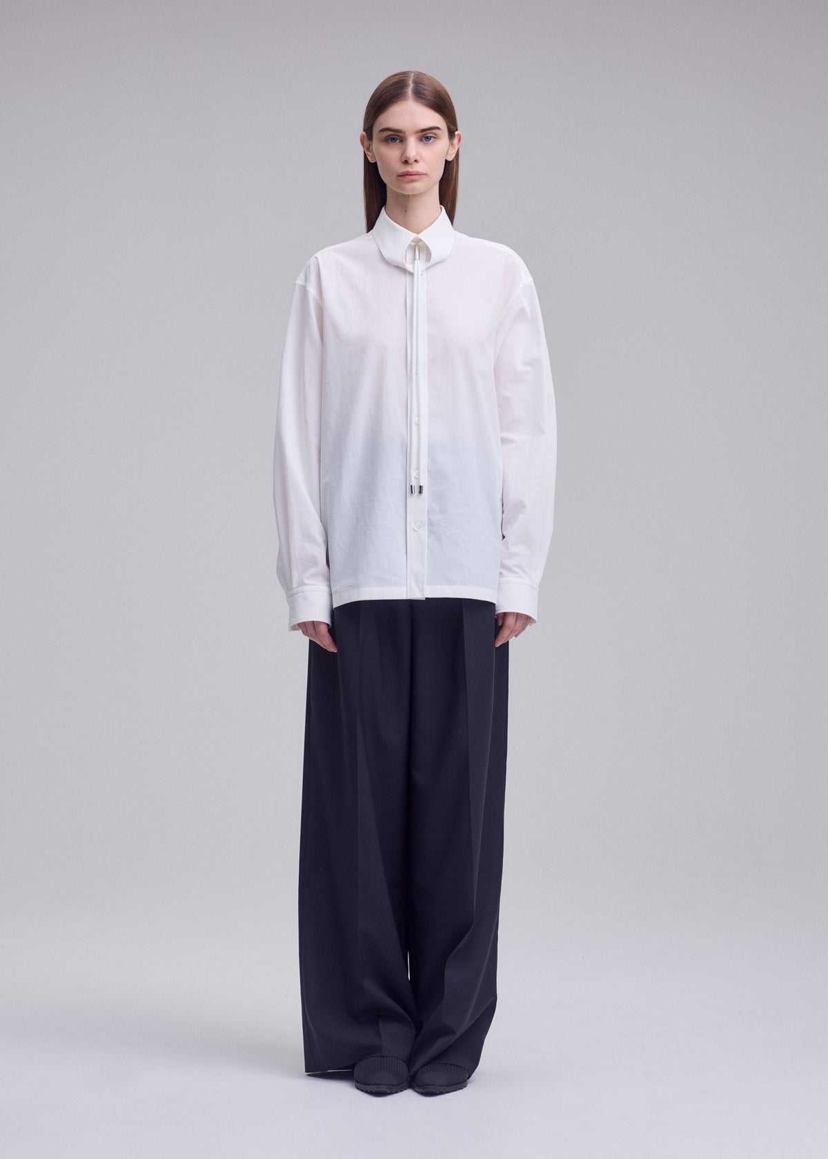 A person stands facing forward, wearing the ISSEY MIYAKE MONSIEUR SHIRT—a loose white high-twist cotton button-up with a string tie—paired with oversized dark wide-leg pants, against a plain light gray background. view 4 of 9