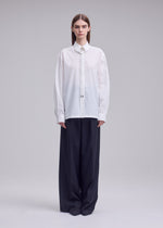 A person stands facing forward, wearing the ISSEY MIYAKE MONSIEUR SHIRT—a loose white high-twist cotton button-up with a string tie—paired with oversized dark wide-leg pants, against a plain light gray background.