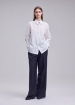 A person stands against a plain background, wearing the ISSEY MIYAKE MONSIEUR SHIRT—a loose high-twist cotton shirt with a regular collar and bolo tie detail—paired with wide-leg black trousers, hands in pockets and looking straight ahead.