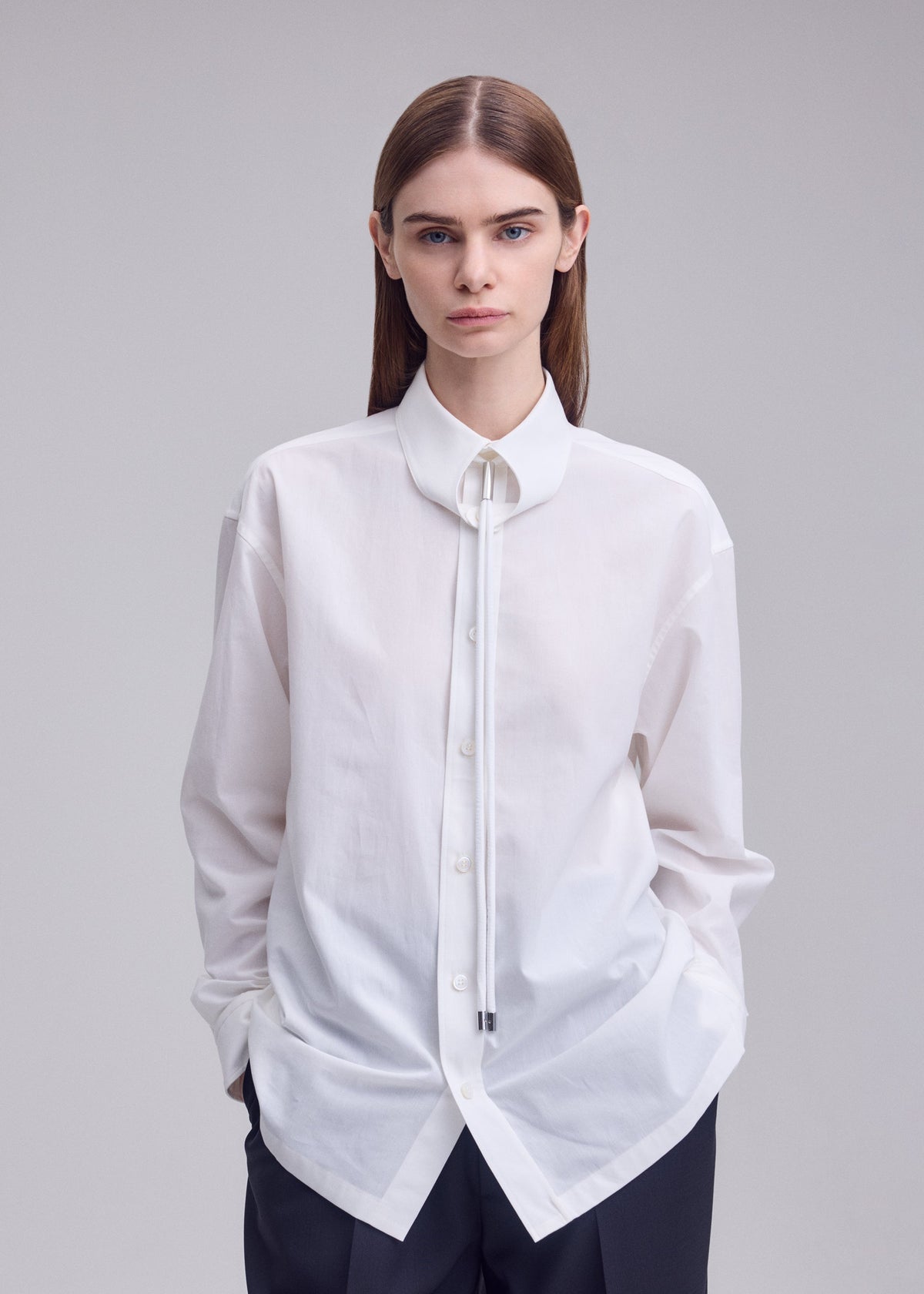 A person with straight brown hair wears the ISSEY MIYAKE MONSIEUR SHIRT, a white regular collar shirt crafted from high-twist cotton, styled with a thin bolo tie around the collar and paired with black pants. They are standing against a plain light gray background. view 6 of 9
