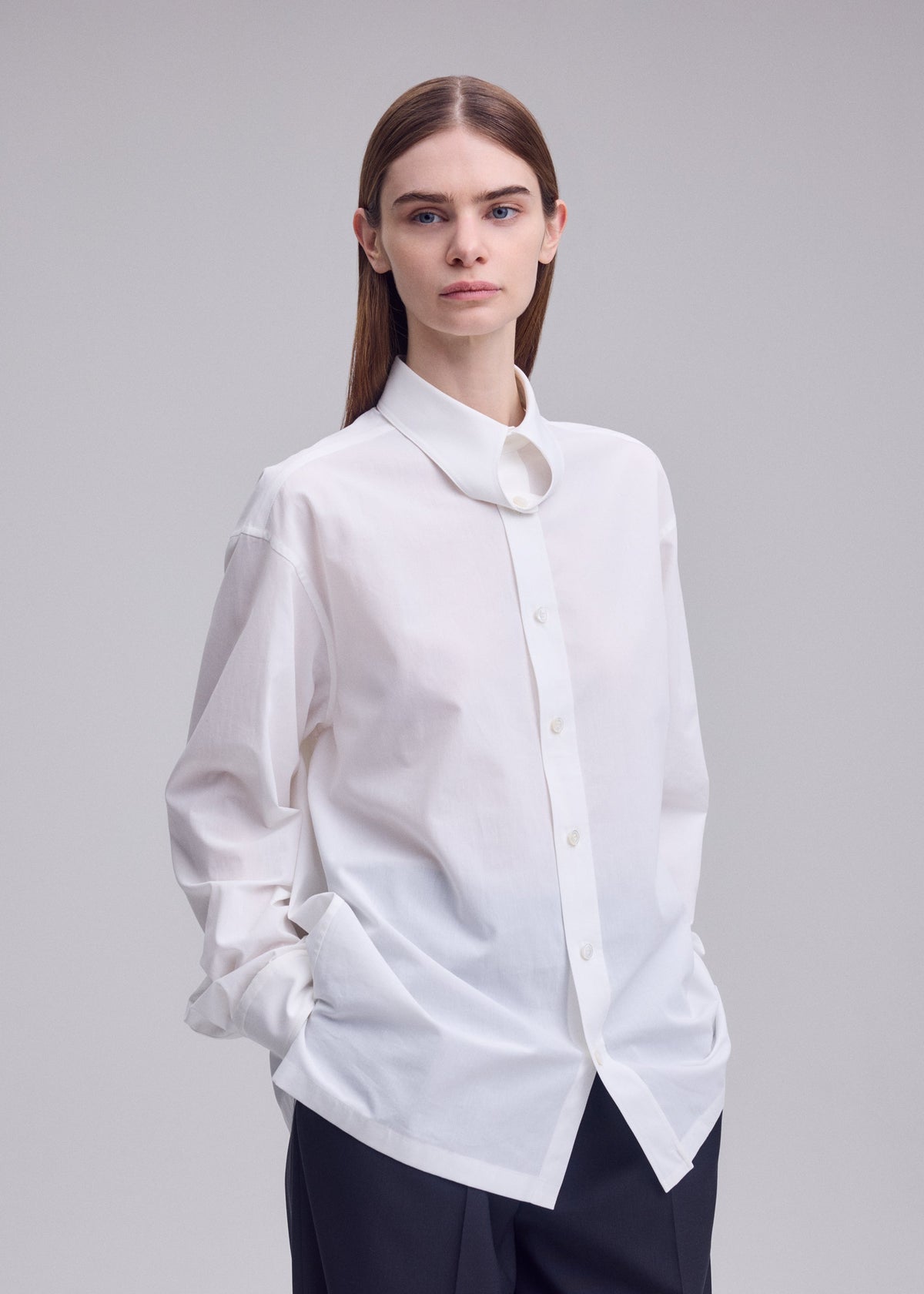 A person with straight brown hair wears the oversized white MONSIEUR SHIRT by ISSEY MIYAKE, crafted from high-twist cotton and featuring a regular collar, paired with dark pants and standing with hands in pockets against a plain light background. view 7 of 9