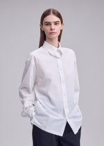 A person with straight brown hair wears the oversized white MONSIEUR SHIRT by ISSEY MIYAKE, crafted from high-twist cotton and featuring a regular collar, paired with dark pants and standing with hands in pockets against a plain light background.