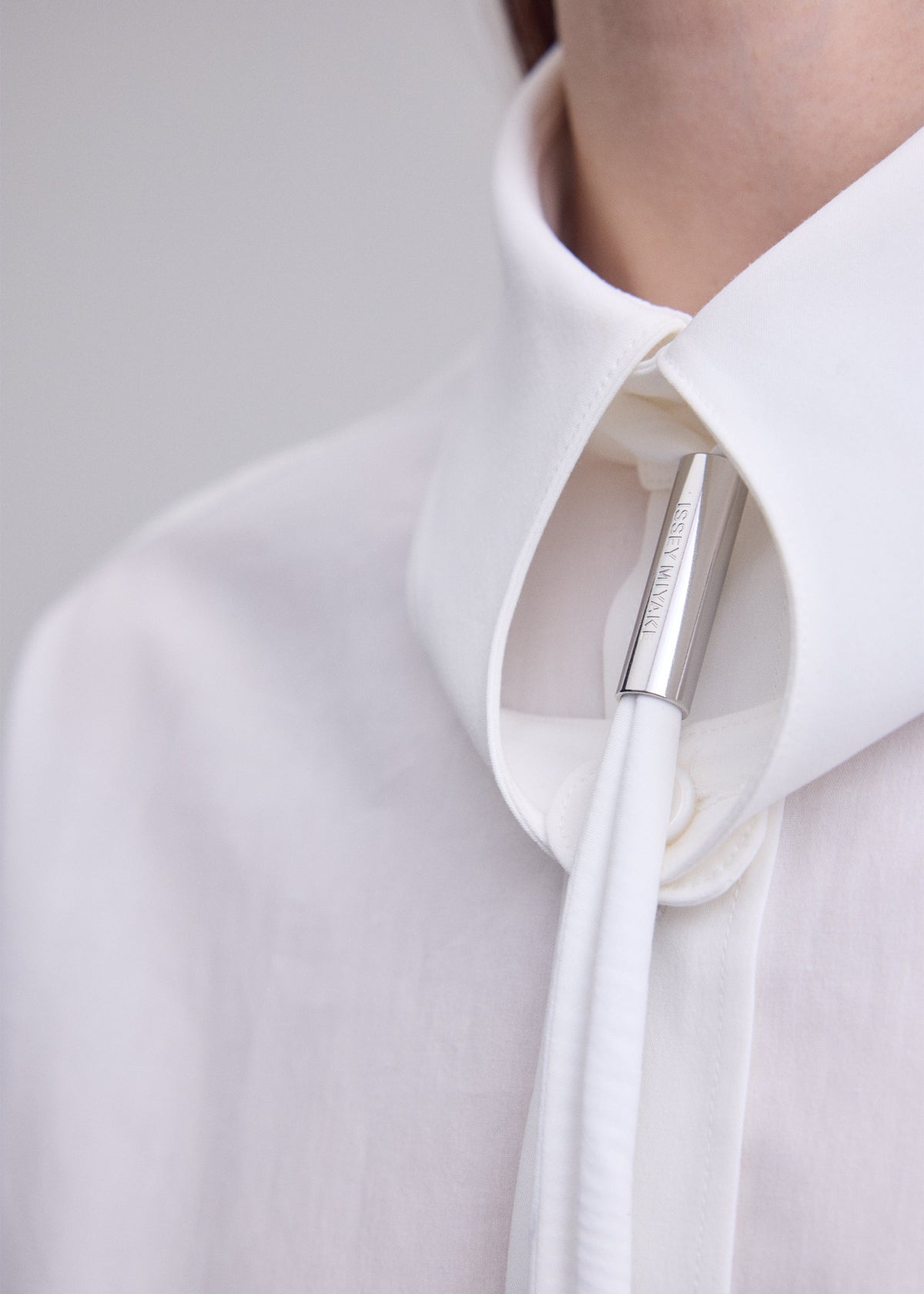 A close-up of a person wearing the ISSEY MIYAKE MONSIEUR SHIRT, featuring a regular collar and crafted from high-twist cotton, paired with a white necktie fastened by a silver cylindrical clasp engraved with small text. The background is plain and light. view 8 of 9