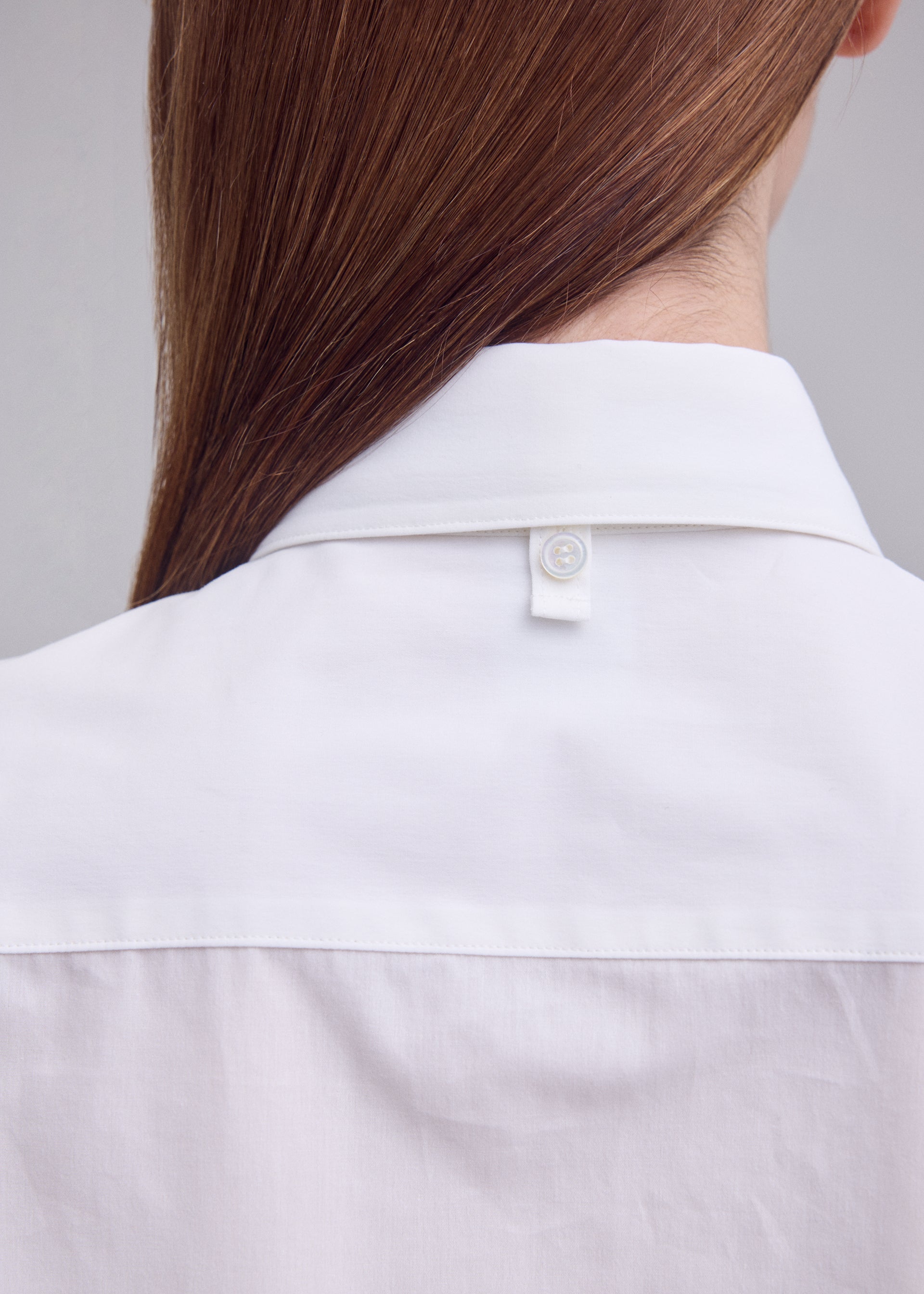 Zoomed view Close-up of the back of a person wearing the MONSIEUR SHIRT by ISSEY MIYAKE, featuring a regular collar and crafted from high-twist cotton, with straight brown hair partially visible and a button detail on the back of the collar.