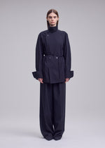 A person stands facing forward wearing the ISSEY MIYAKE MONSIEUR SHIRT, a dark, high-collared belted haori-style jacket with large cuffs, paired with wide-leg trousers against a neutral background.