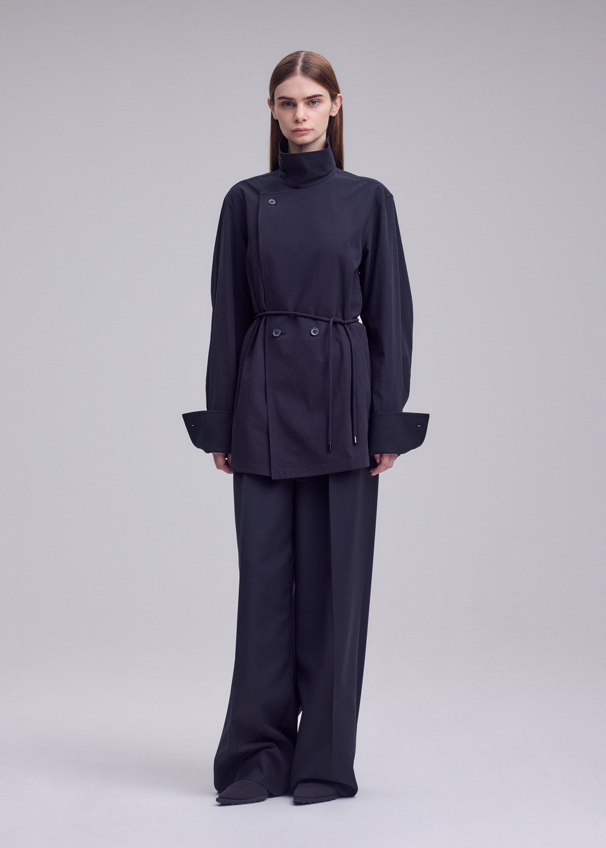 A person stands against a plain background, wearing the ISSEY MIYAKE MONSIEUR SHIRT—a dark, belted long-sleeve tunic—paired with wide-leg trousers and a bolo tie at the collar. Their hair is straight and parted in the middle, enhancing the ensemble’s minimalist and modern aesthetic. view 5 of 7