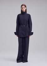 A person stands against a plain background, wearing the ISSEY MIYAKE MONSIEUR SHIRT—a dark, belted long-sleeve tunic—paired with wide-leg trousers and a bolo tie at the collar. Their hair is straight and parted in the middle, enhancing the ensemble’s minimalist and modern aesthetic.