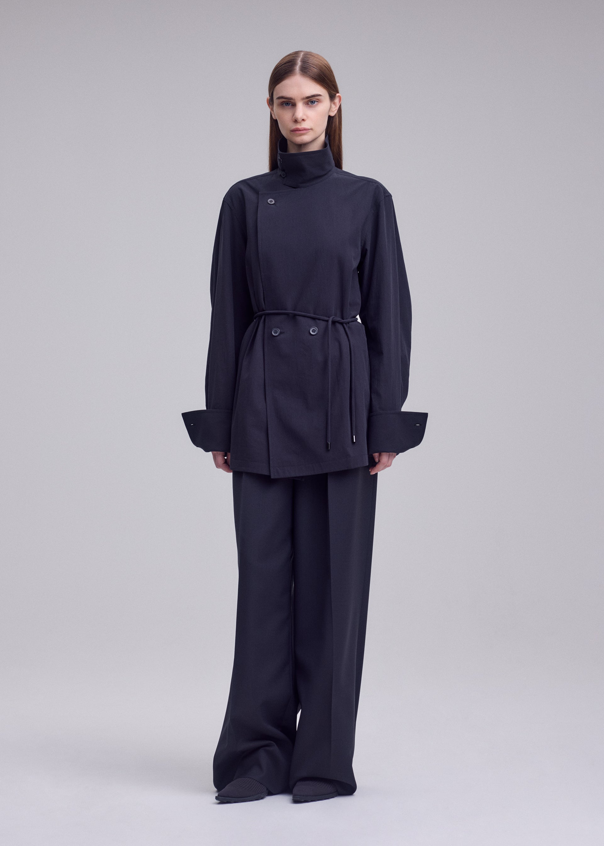 Zoomed view A person stands against a plain background, wearing the ISSEY MIYAKE MONSIEUR SHIRT—a dark, belted long-sleeve tunic—paired with wide-leg trousers and a bolo tie at the collar. Their hair is straight and parted in the middle, enhancing the ensemble’s minimalist and modern aesthetic.