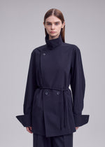 A person stands against a plain background, wearing the MONSIEUR SHIRT by ISSEY MIYAKE—a dark, long-sleeved asymmetric haori-style jacket featuring a high collar, wide cuffs, button details, and a thin waist belt.