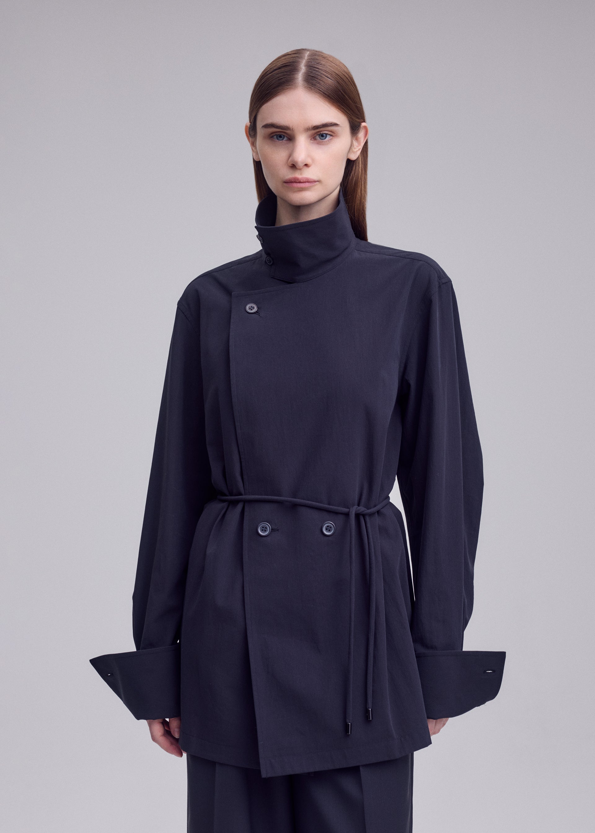 Zoomed view A person stands against a plain background, wearing the MONSIEUR SHIRT by ISSEY MIYAKE—a dark, long-sleeved asymmetric haori-style jacket featuring a high collar, wide cuffs, button details, and a thin waist belt.