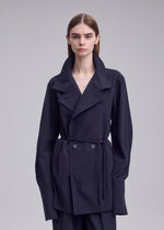 A person with straight brown hair stands against a plain light gray background, wearing the ISSEY MIYAKE MONSIEUR SHIRT beneath a belted, oversized black trench coat featuring wide lapels and long sleeves.