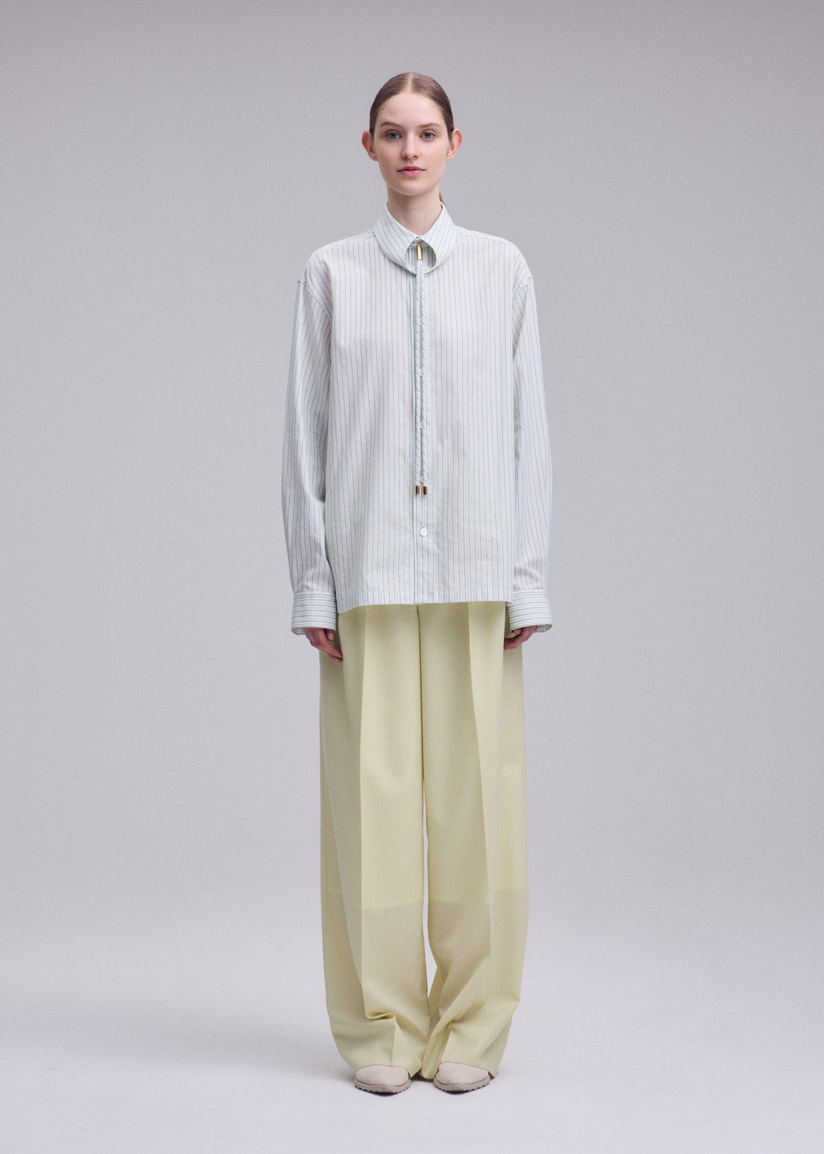 A person stands facing forward against a plain light background, wearing ISSEY MIYAKE’s MONSIEUR SHIRT (DOBBY STRIPE), an oversized light blue shirt crafted from cotton yarn-dyed fabric, paired with wide-legged beige trousers and light shoes. view 5 of 10