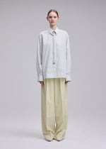 A person stands facing forward against a plain light background, wearing ISSEY MIYAKE’s MONSIEUR SHIRT (DOBBY STRIPE), an oversized light blue shirt crafted from cotton yarn-dyed fabric, paired with wide-legged beige trousers and light shoes.