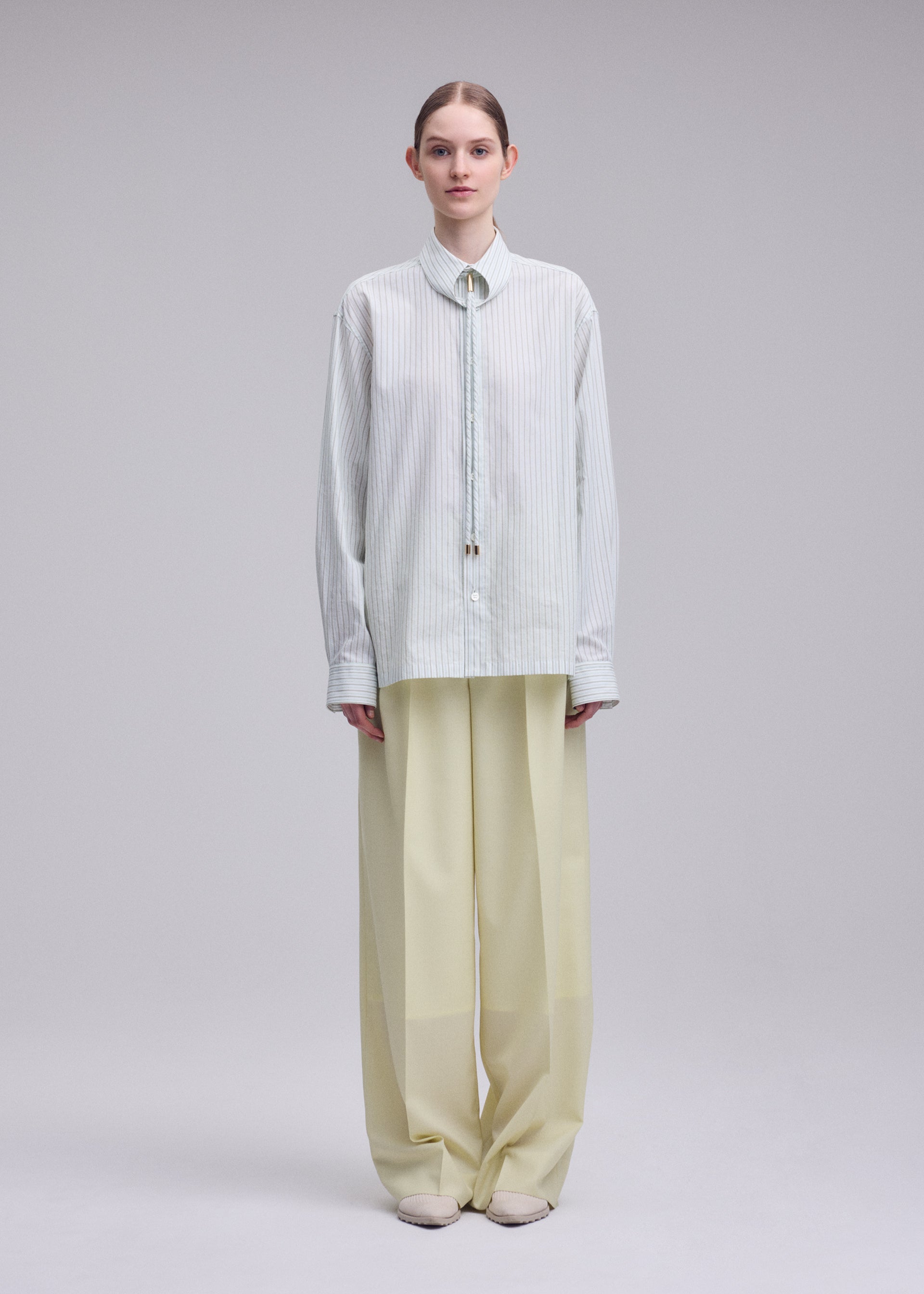 Zoomed view A person stands facing forward against a plain light background, wearing ISSEY MIYAKE’s MONSIEUR SHIRT (DOBBY STRIPE), an oversized light blue shirt crafted from cotton yarn-dyed fabric, paired with wide-legged beige trousers and light shoes.