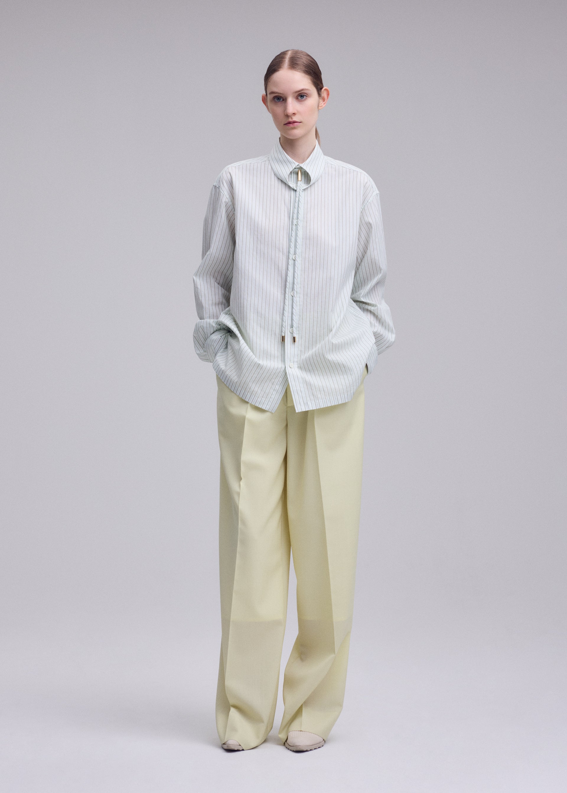 Zoomed view Against a plain background, a person stands with hands in pockets, wearing the ISSEY MIYAKE MONSIEUR SHIRT (DOBBY STRIPE)—a light striped button-up—and loose, cream-colored trousers; their hair is neatly pulled back.