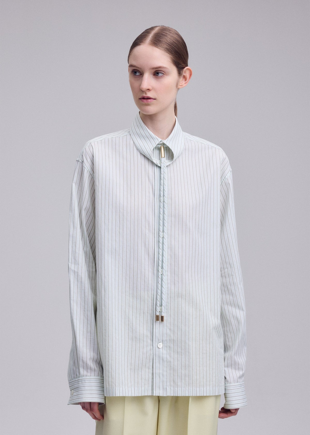 A person with light skin and brown hair styled in a low bun wears the ISSEY MIYAKE MONSIEUR SHIRT(DOBBY STRIPE), an oversized shirt made from cotton yarn-dyed dobby stripe fabric, featuring a narrow collar and paired with a bolo tie and light-colored pants, standing against a plain background. view 7 of 10