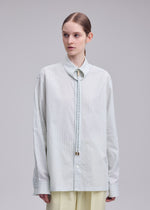 A person with light skin and brown hair styled in a low bun wears the ISSEY MIYAKE MONSIEUR SHIRT(DOBBY STRIPE), an oversized shirt made from cotton yarn-dyed dobby stripe fabric, featuring a narrow collar and paired with a bolo tie and light-colored pants, standing against a plain background.
