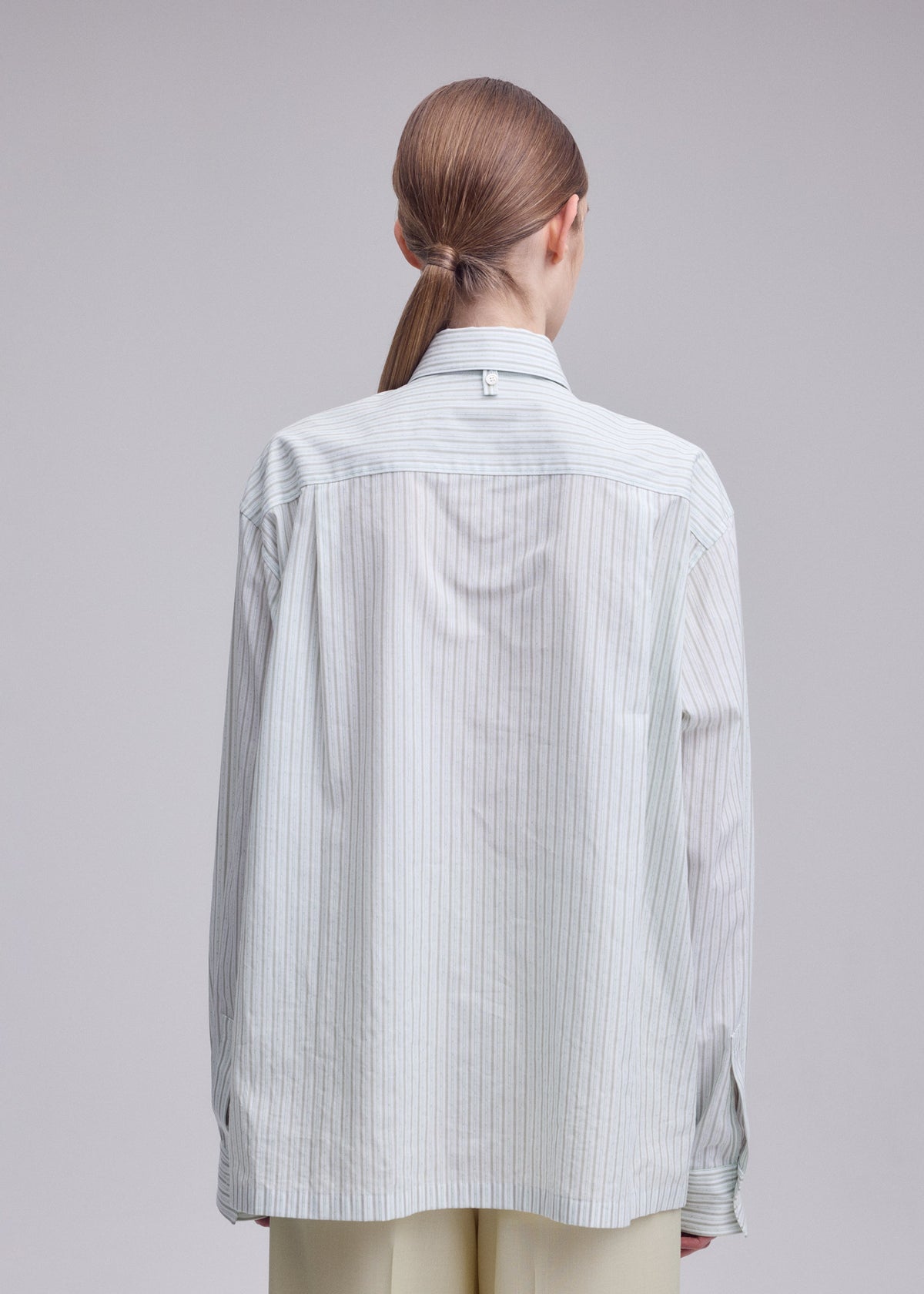 A person with straight, light brown hair in a low ponytail is shown from the back, wearing the ISSEY MIYAKE MONSIEUR SHIRT (DOBBY STRIPE), an oversized shirt crafted from cotton yarn-dyed fabric, paired with light trousers against a plain background. view 8 of 10