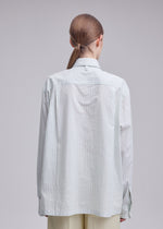 A person with straight, light brown hair in a low ponytail is shown from the back, wearing the ISSEY MIYAKE MONSIEUR SHIRT (DOBBY STRIPE), an oversized shirt crafted from cotton yarn-dyed fabric, paired with light trousers against a plain background.