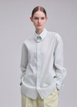 A person wearing the ISSEY MIYAKE MONSIEUR SHIRT (DOBBY STRIPE)—a light-colored, loose-fitting shirt crafted from cotton yarn-dyed fabric—paired with light pants, stands against a plain background with hands in pockets, gazing slightly to the side.