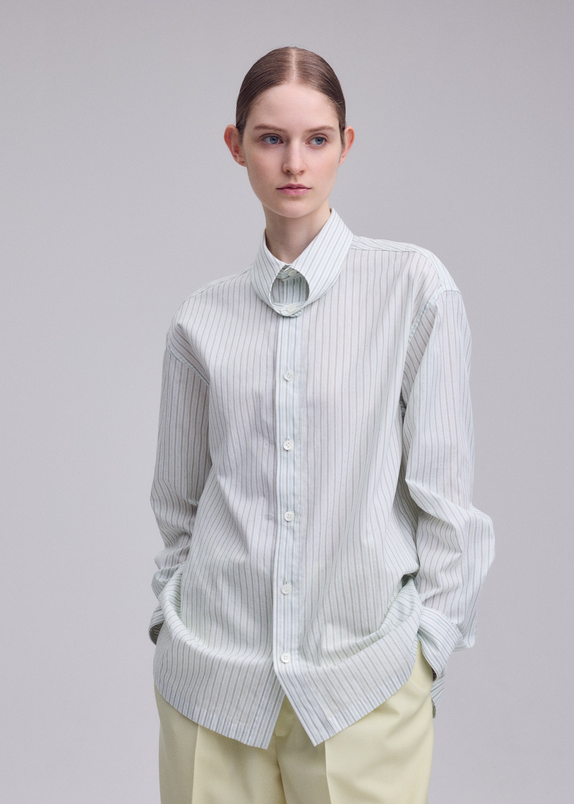 Zoomed view A person wearing the ISSEY MIYAKE MONSIEUR SHIRT (DOBBY STRIPE)—a light-colored, loose-fitting shirt crafted from cotton yarn-dyed fabric—paired with light pants, stands against a plain background with hands in pockets, gazing slightly to the side.