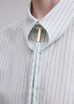 A close-up of a person wearing the ISSEY MIYAKE MONSIEUR SHIRT (DOBBY STRIPE), crafted from cotton yarn-dyed fabric, paired with a bolo tie featuring a gold clasp.