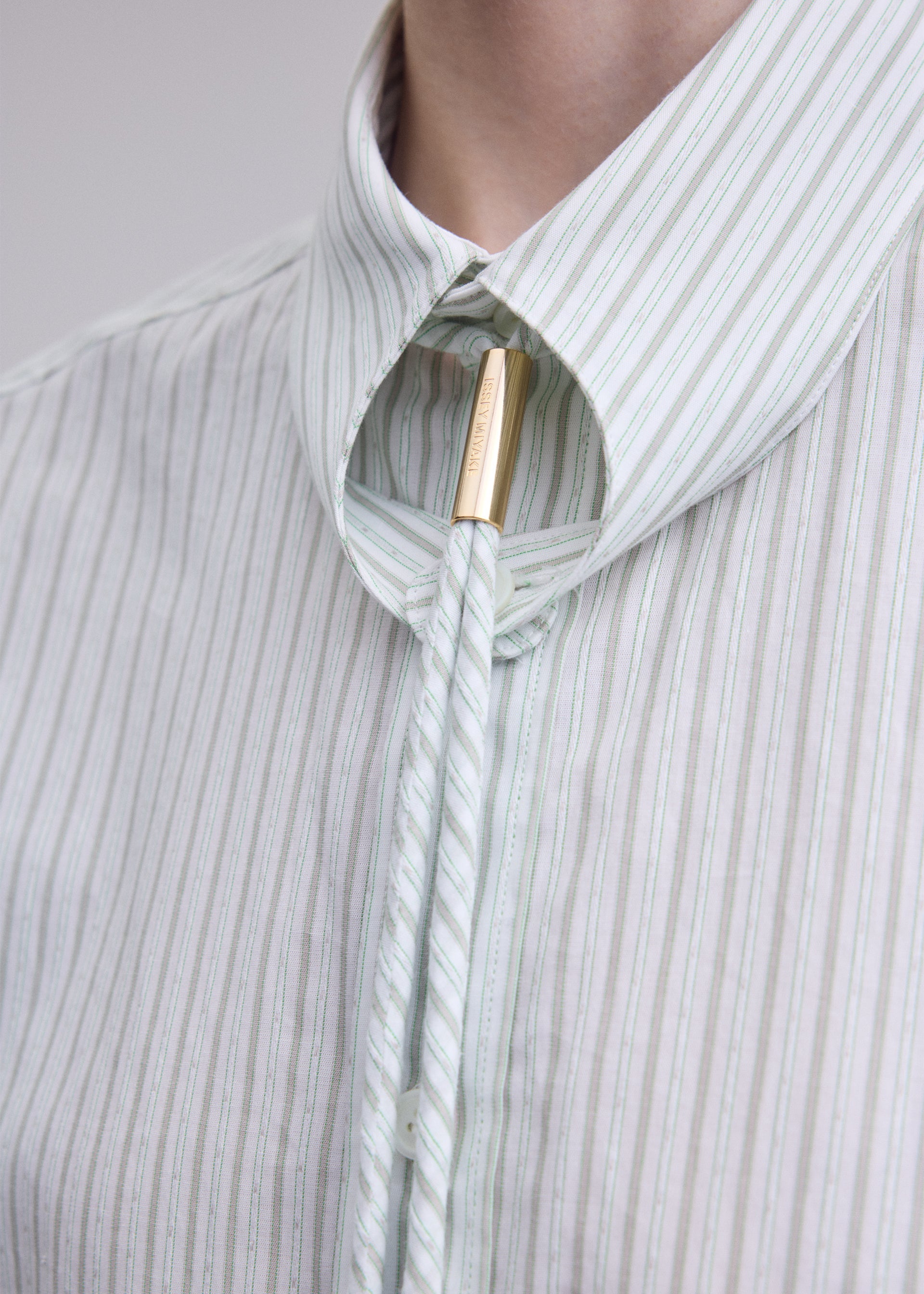 Zoomed view A close-up of a person wearing the ISSEY MIYAKE MONSIEUR SHIRT (DOBBY STRIPE), crafted from cotton yarn-dyed fabric, paired with a bolo tie featuring a gold clasp.
