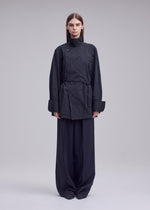 A person faces forward against a plain light background, wearing the ISSEY MIYAKE MONSIEUR SHIRT (DOBBY STRIPE), a dark, pinstriped jacket-style shirt crafted from cotton yarn-dyed fabric with wide sleeves, paired with loose, wide-leg trousers.