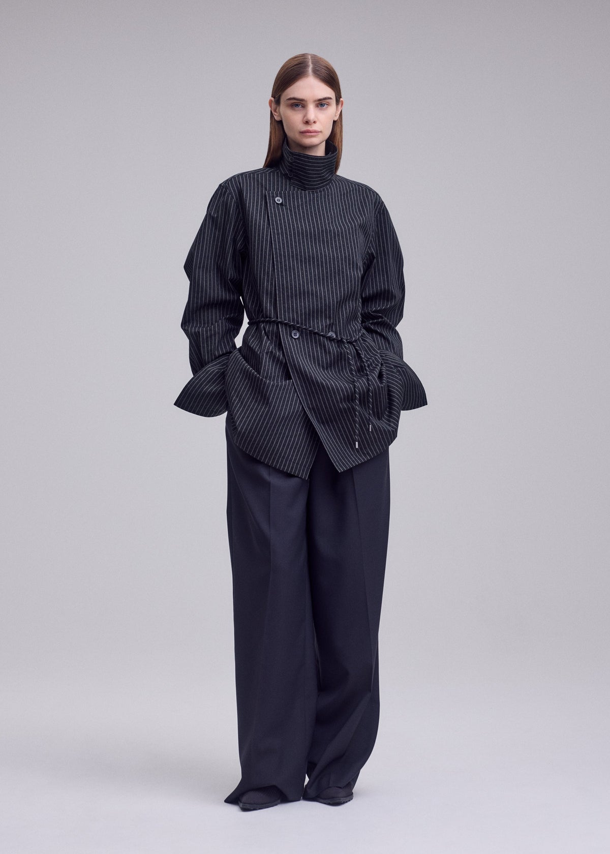 A woman stands wearing the ISSEY MIYAKE MONSIEUR SHIRT (DOBBY STRIPE), featuring a dark yarn-dyed pinstripe design. She pairs it with wide-leg trousers, her straight brown hair and neutral expression complementing the minimal, light gray background. view 6 of 8
