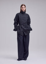 A woman stands wearing the ISSEY MIYAKE MONSIEUR SHIRT (DOBBY STRIPE), featuring a dark yarn-dyed pinstripe design. She pairs it with wide-leg trousers, her straight brown hair and neutral expression complementing the minimal, light gray background.
