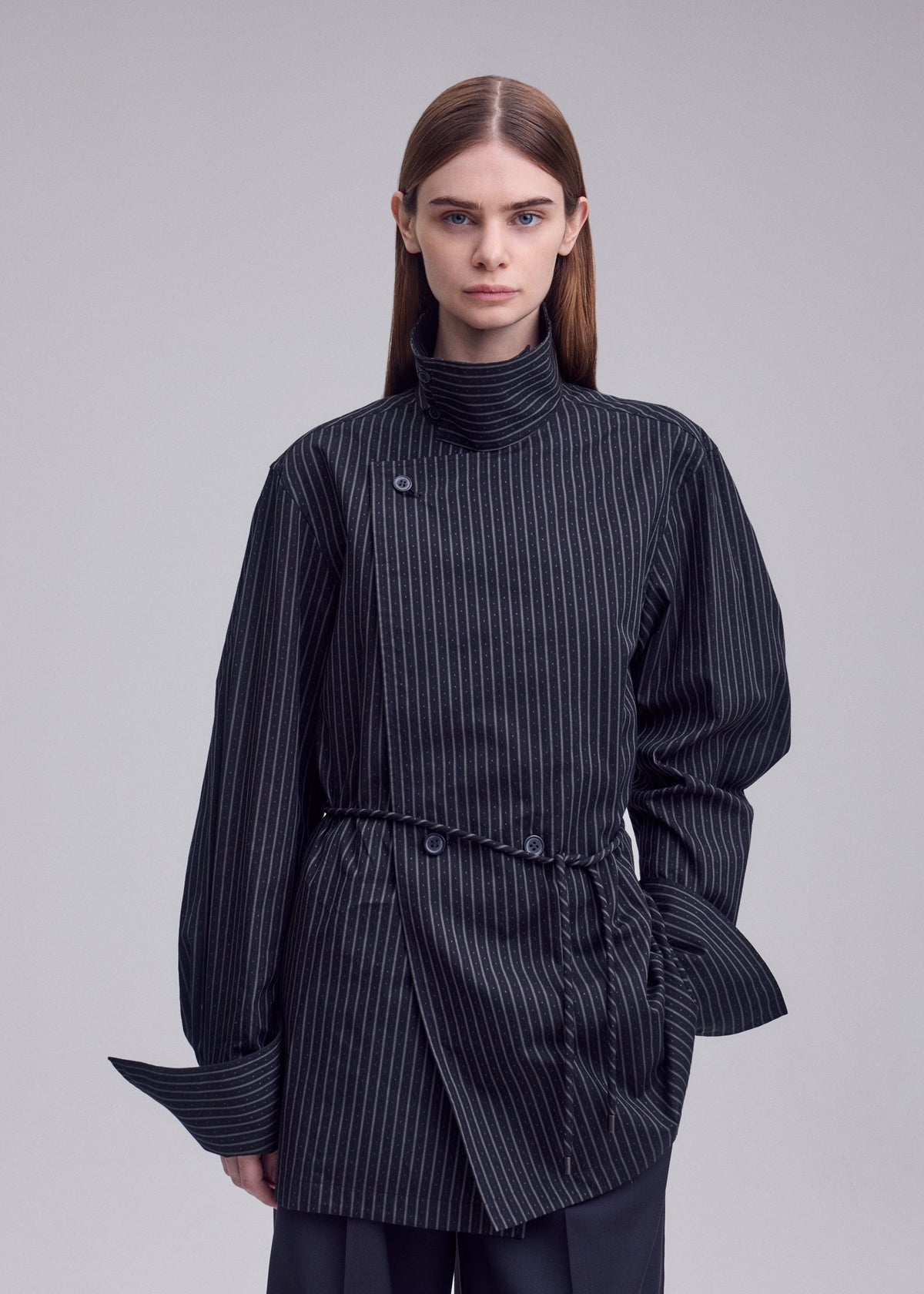 A person with straight brown hair wears the ISSEY MIYAKE MONSIEUR SHIRT (DOBBY STRIPE), a structured black top featuring pinstripes, a high collar, and unique button details. The background is plain and light gray. view 7 of 8
