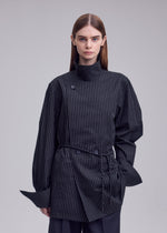 A person with straight brown hair wears the ISSEY MIYAKE MONSIEUR SHIRT (DOBBY STRIPE), a structured black top featuring pinstripes, a high collar, and unique button details. The background is plain and light gray.