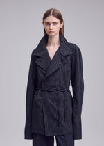 A person stands against a plain gray background, looking straight ahead with a neutral expression, dressed in a dark, pinstriped jacket featuring wide lapels, oversized cuffs, and a tie belt. Under the jacket, they wear the ISSEY MIYAKE MONSIEUR SHIRT (DOBBY STRIPE), crafted from soft cotton yarn-dyed fabric that subtly adds texture to the ensemble.