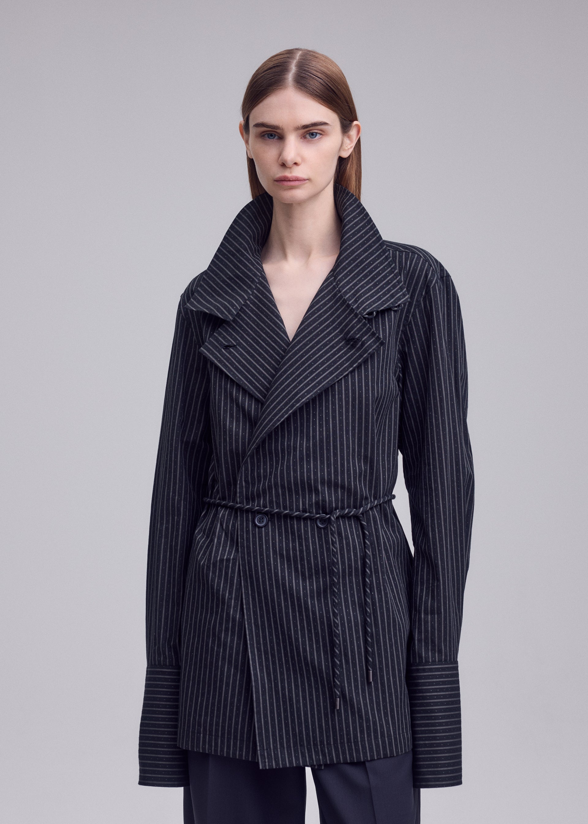 Zoomed view A person stands against a plain gray background, looking straight ahead with a neutral expression, dressed in a dark, pinstriped jacket featuring wide lapels, oversized cuffs, and a tie belt. Under the jacket, they wear the ISSEY MIYAKE MONSIEUR SHIRT (DOBBY STRIPE), crafted from soft cotton yarn-dyed fabric that subtly adds texture to the ensemble.