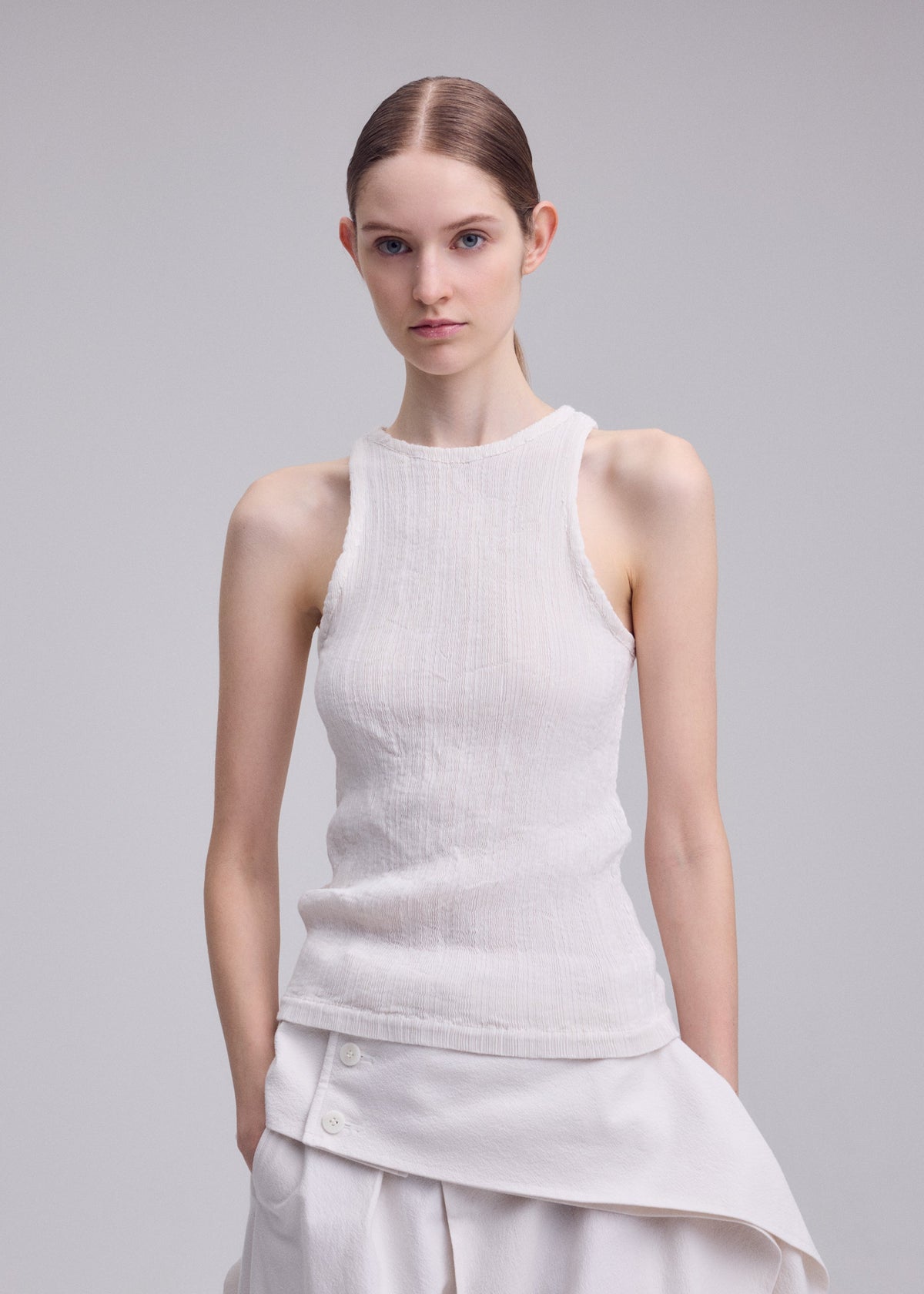 A young woman with fair skin and straight brown hair, parted in the middle, models the ISSEY MIYAKE RILL round-neck sleeveless top and white skirt with button detailing, both featuring subtly pleated fabric, against a light gray background. view 5 of 6