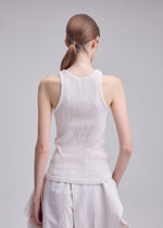 A person with straight, light brown hair in a low ponytail stands with their back to the camera, wearing ISSEY MIYAKE's RILL round-neck sleeveless top and loose pleated white pants against a plain light gray background.