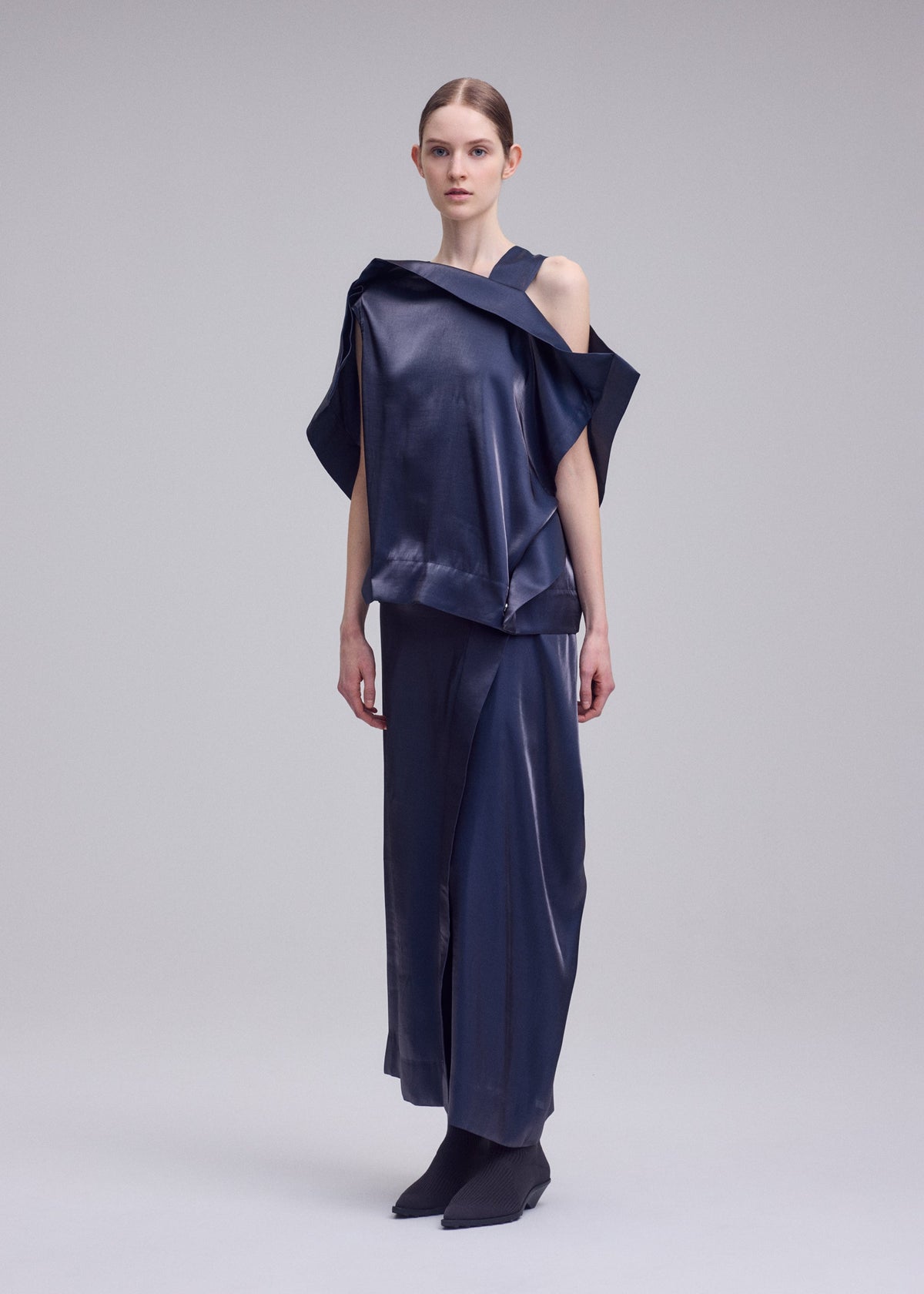 A woman stands against a plain background wearing the MERCURY top and matching long skirt by ISSEY MIYAKE—an asymmetrical navy Italian cloth set with a subtle metallic sheen, one shoulder exposed. Her straight hair is pulled back and she's in black shoes. view 6 of 7