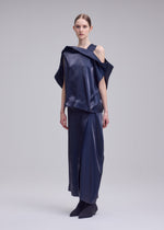 A woman stands against a plain background wearing the MERCURY top and matching long skirt by ISSEY MIYAKE—an asymmetrical navy Italian cloth set with a subtle metallic sheen, one shoulder exposed. Her straight hair is pulled back and she's in black shoes.