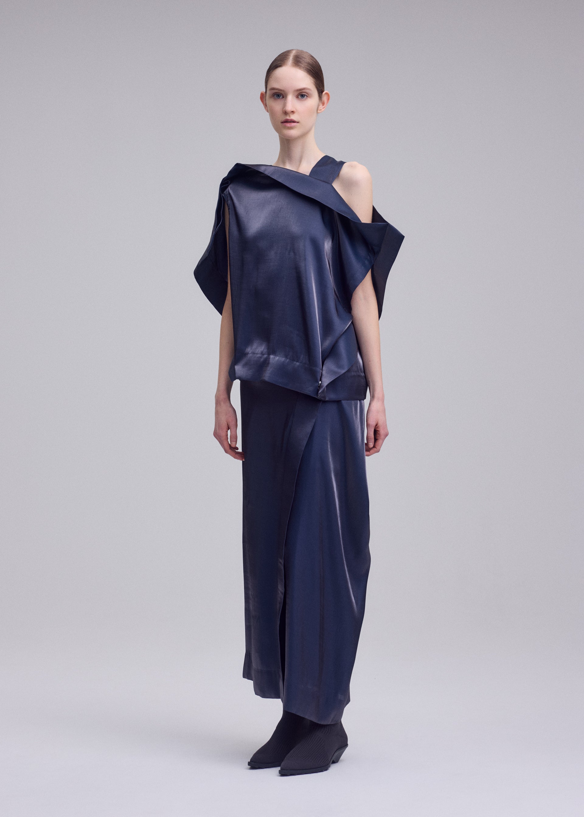 Zoomed view A woman stands against a plain background wearing the MERCURY top and matching long skirt by ISSEY MIYAKE—an asymmetrical navy Italian cloth set with a subtle metallic sheen, one shoulder exposed. Her straight hair is pulled back and she's in black shoes.